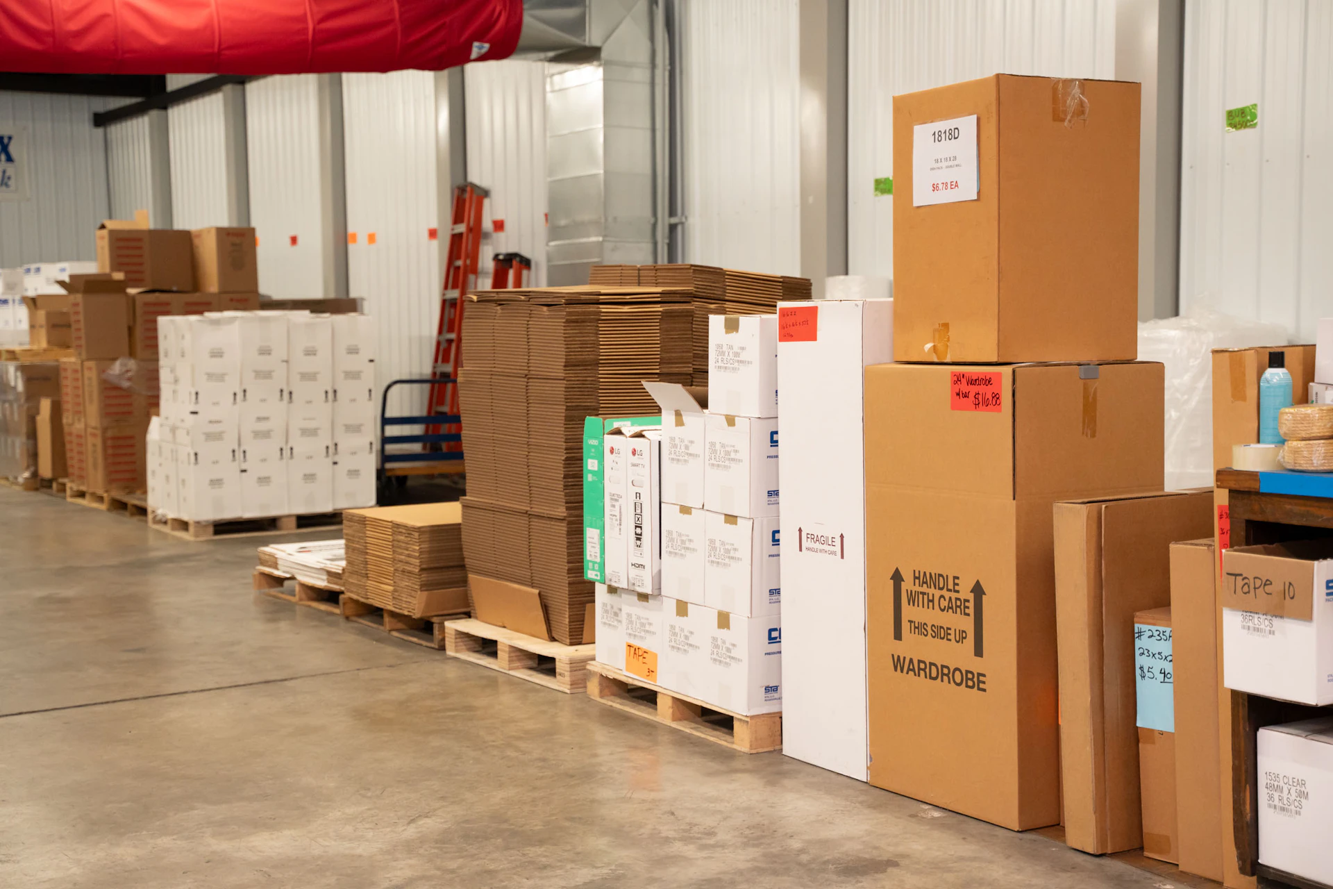 Stacks of cardboard boxes and packing materials are organized on pallets inside a warehouse with concrete floors and metal walls. Some boxes are labeled “fragile” and “wardrobe.” A red ladder is visible in the background.