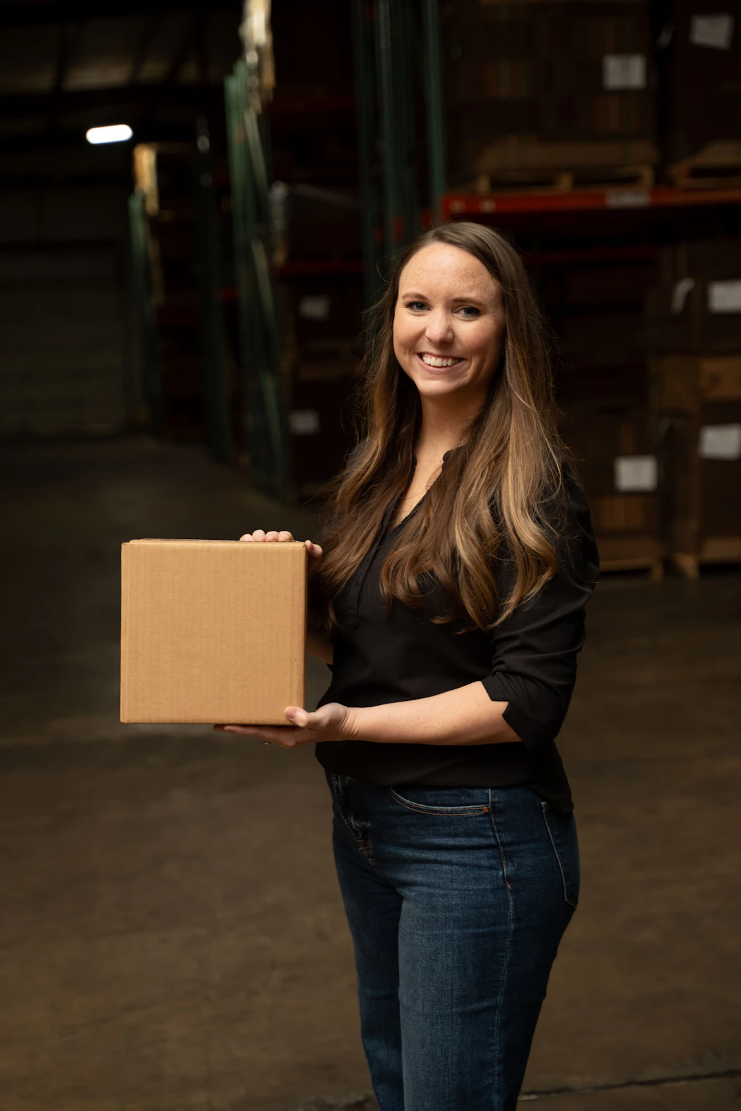 A woman with long brown hair, wearing a black shirt and jeans, stands in a warehouse and smiles while holding a cardboard box.