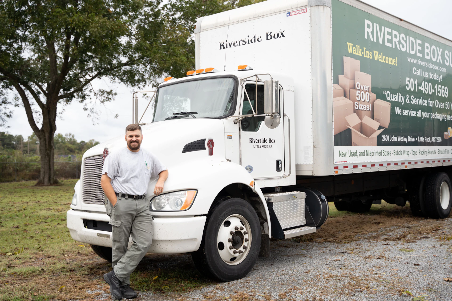 A man in a gray shirt and cargo pants leans against a white Riverside Box moving truck parked outdoors on a gravel surface with grass and trees in the background.