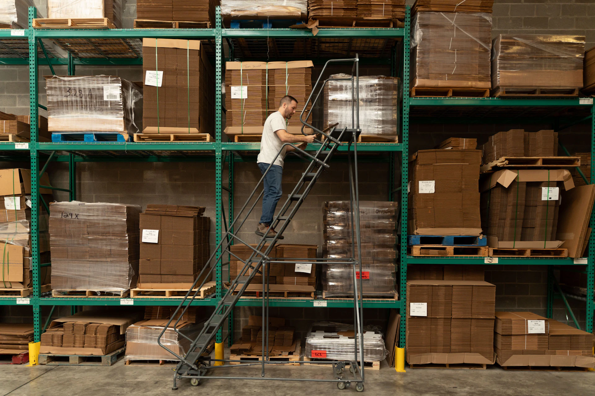 A man stands on a rolling metal ladder, reaching for cardboard boxes on a high warehouse shelf surrounded by neatly stacked pallets and packaging materials.