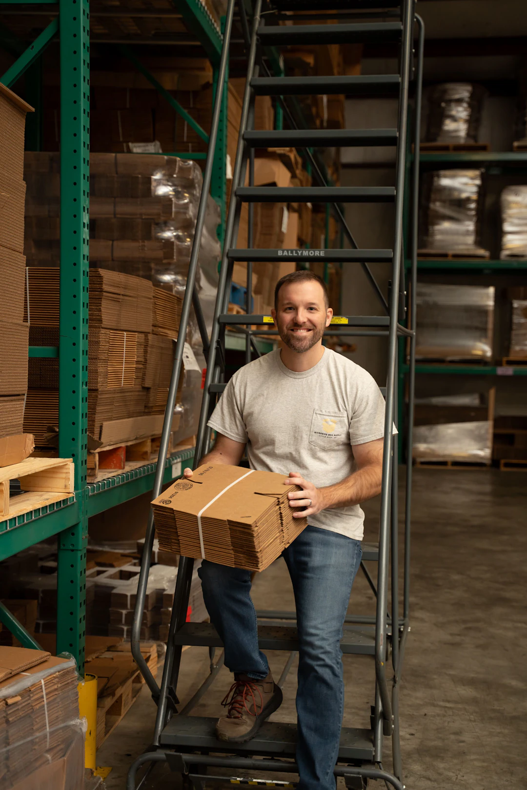 A man in a gray t-shirt and jeans stands on a warehouse ladder, smiling and holding a bundle of flat cardboard boxes. Shelves with stacked boxes and pallets are visible in the background.
