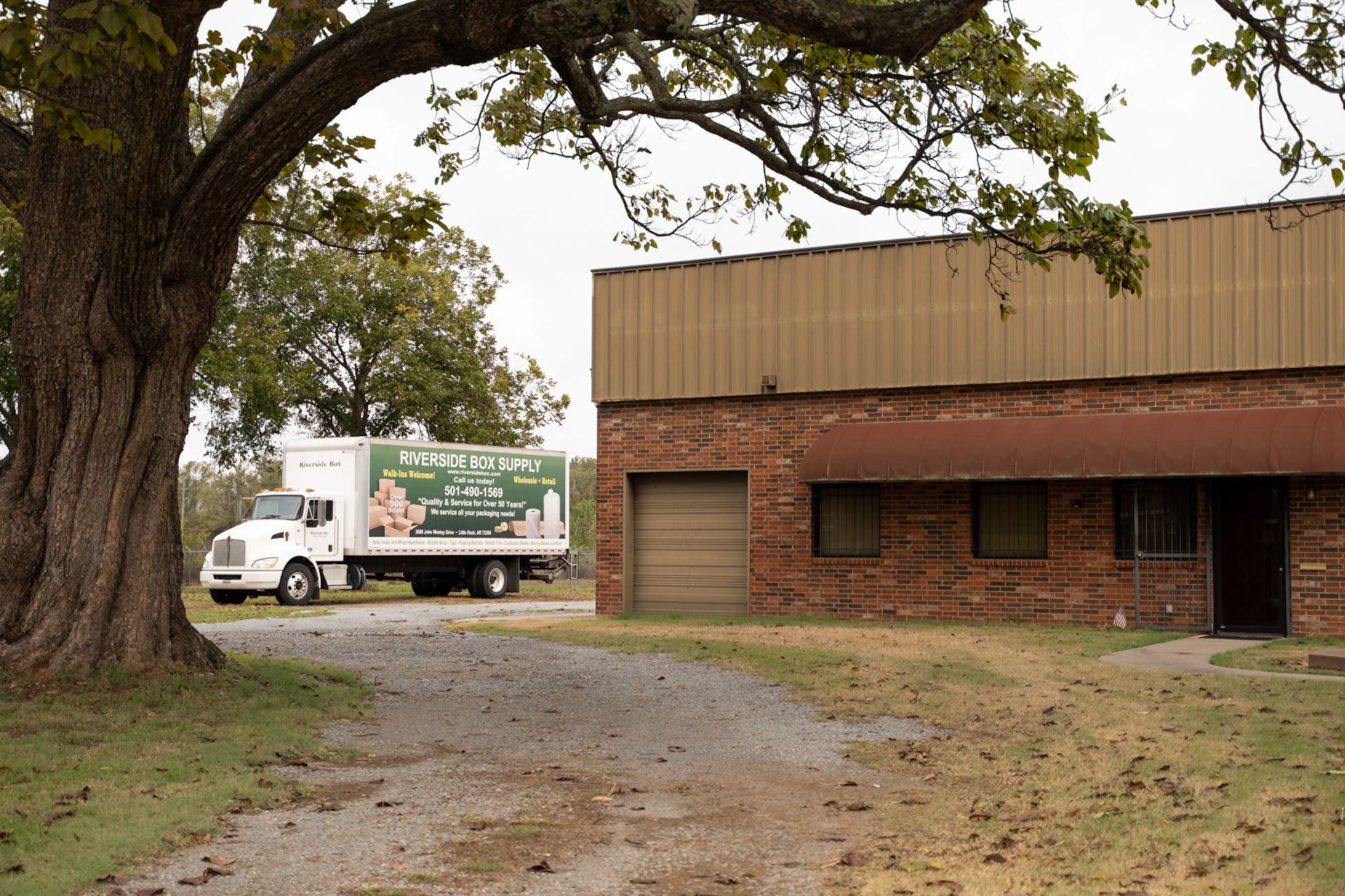 A white Riverside Bolt Supply truck is parked near a brick building with a brown awning, next to a large tree and a gravel driveway on an overcast day.