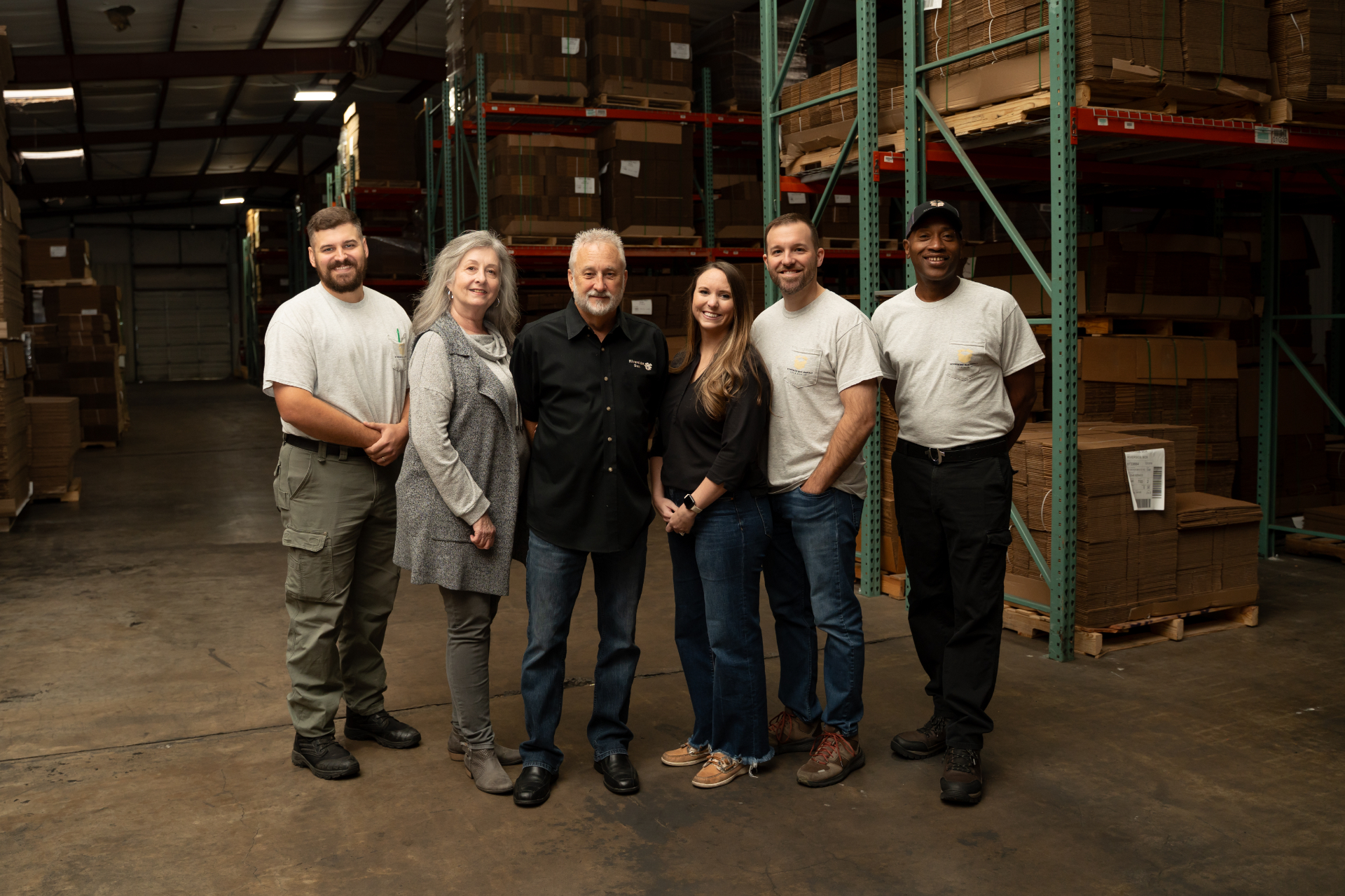 Six people stand together and smile for a group photo in a warehouse, with shelves full of cardboard boxes in the background. They appear to be workers or staff members.
