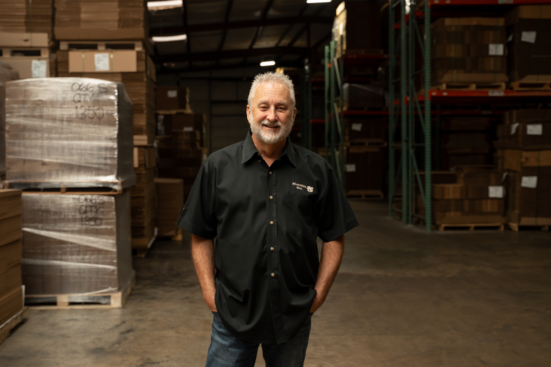 An older man with gray hair and a beard stands smiling in a warehouse, wearing a black short-sleeve button-up shirt and jeans, with stacked boxes and shelves in the background.