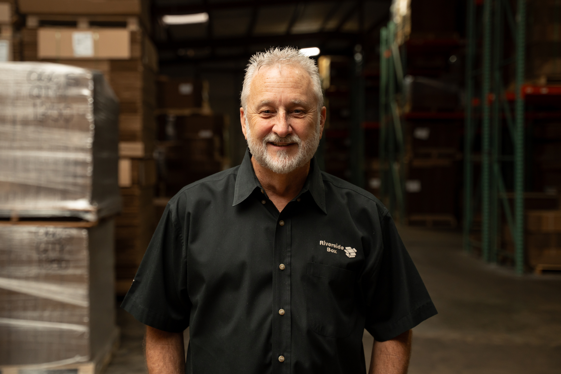 An older man with short gray hair and a beard smiles, standing in a warehouse filled with stacked boxes. He wears a black button-up shirt with a company logo on the chest.