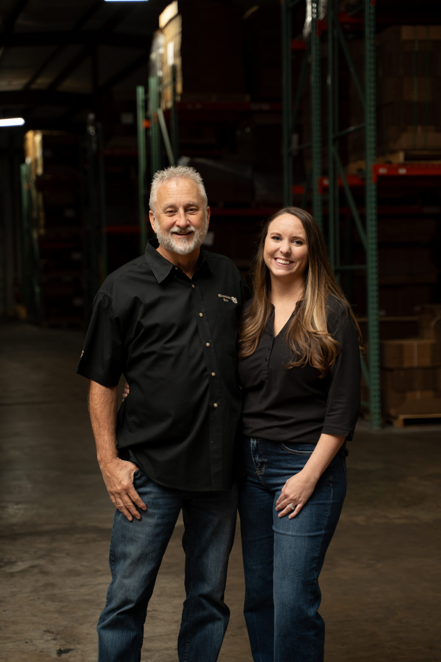 A man and woman stand side by side, smiling, in a warehouse with shelves and boxes in the background. Both are wearing dark shirts and jeans. The lighting is warm, highlighting them against the industrial setting.