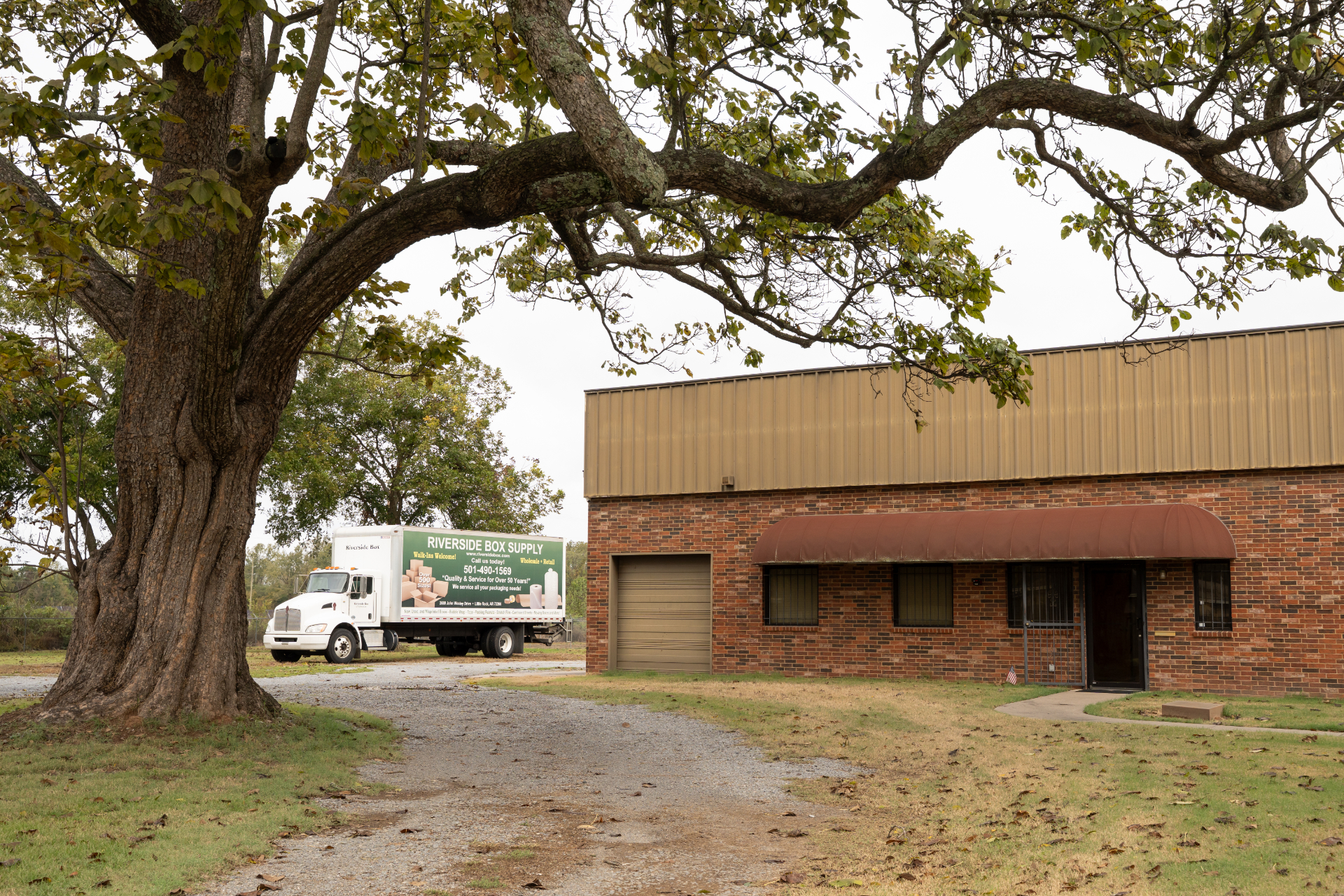 A white truck with “Riverside Bio Supply” and plant images parked beside a brick industrial building with a brown metal roof, large tree in the foreground, and overcast sky.