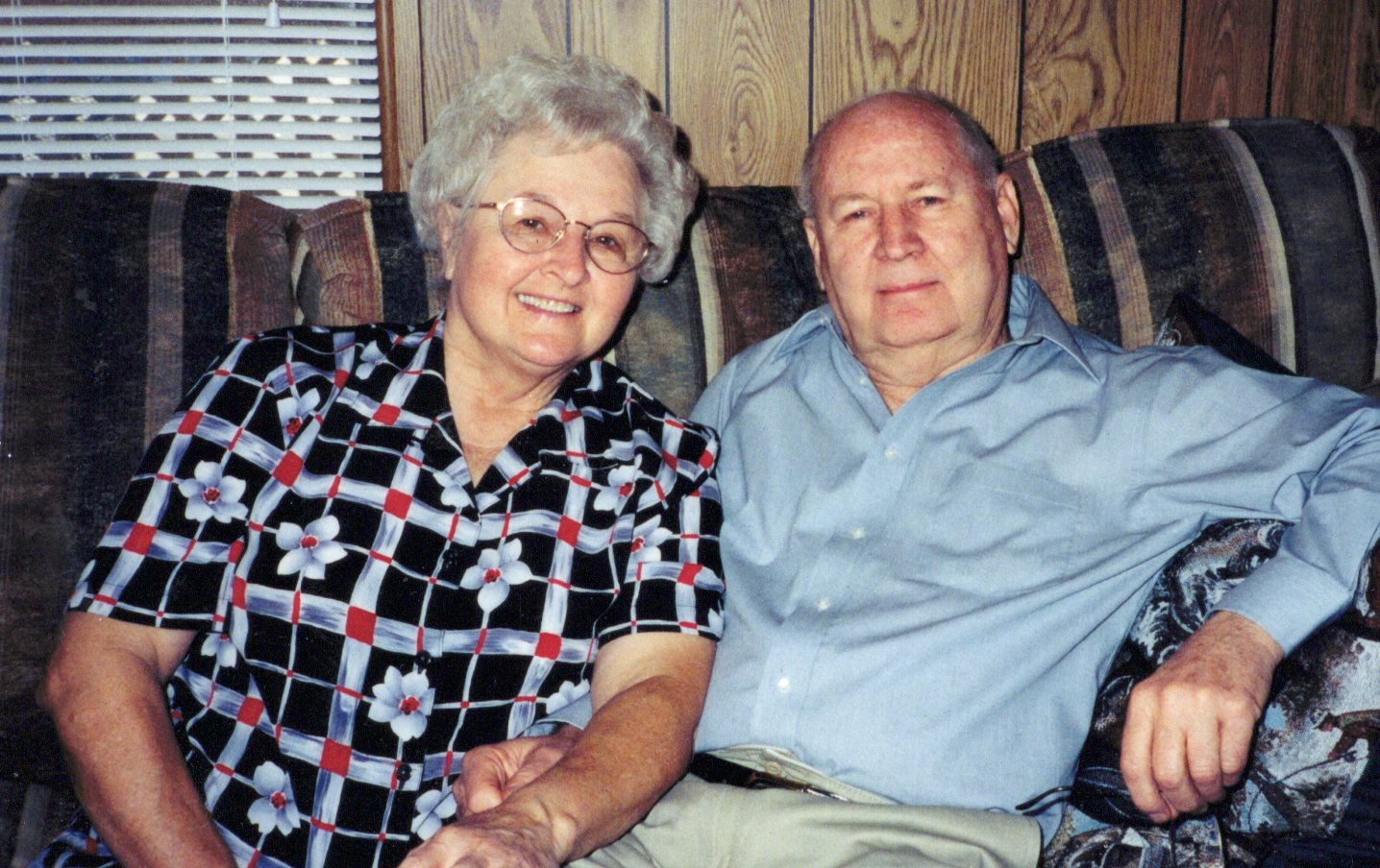An elderly woman and man sit close together on a patterned couch, smiling at the camera. The woman wears glasses and a patterned blouse; the man wears a light blue shirt. Wood paneling and a window are in the background.