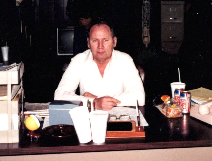 A man in a white shirt sits at a desk with paperwork, two white cups, a can of Pepsi, a sandwich, and various office supplies. File organizers and a rotary phone are also on the desk; a filing cabinet is in the background.