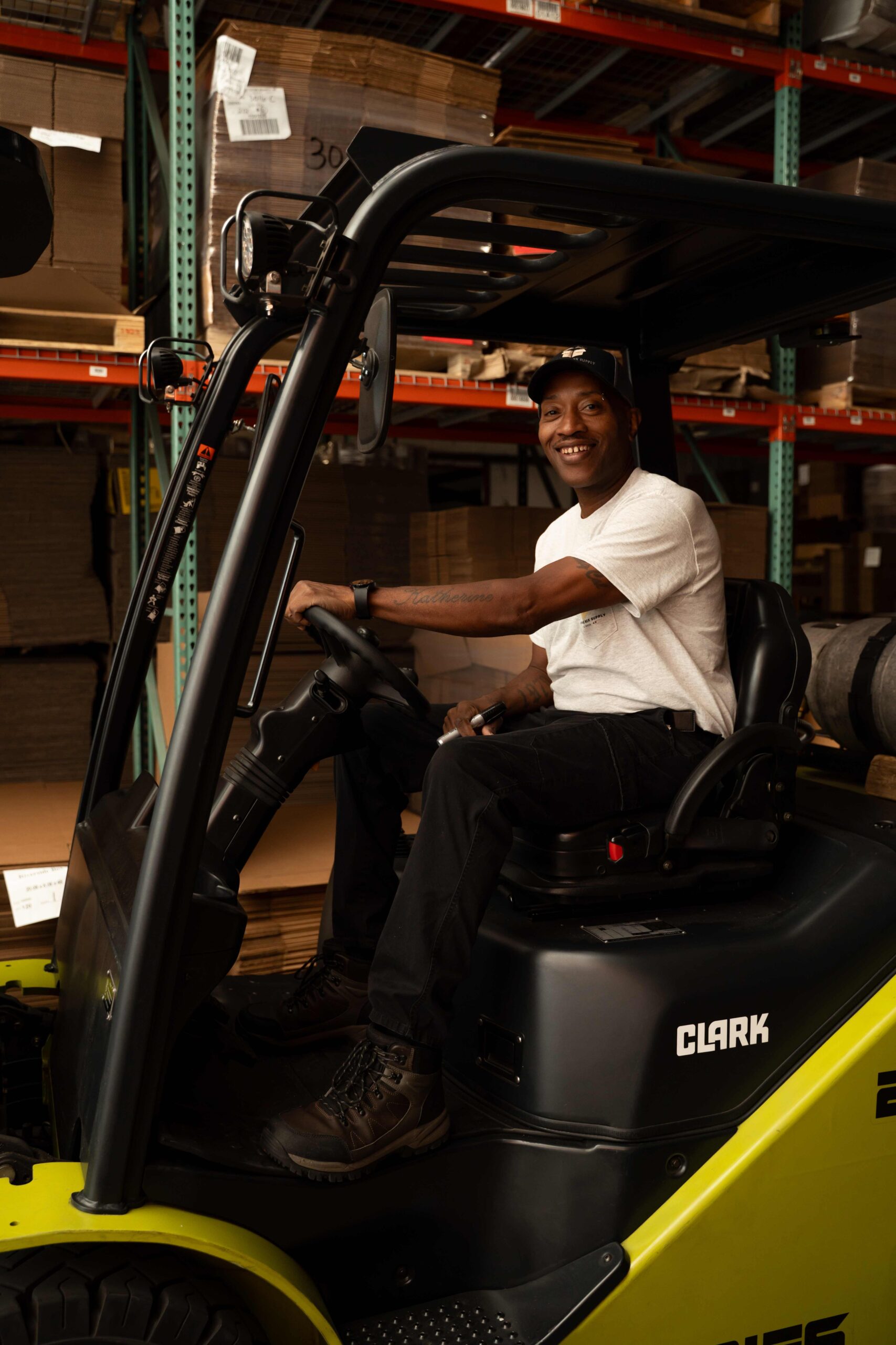 A man wearing a white t-shirt and black pants smiles while sitting on a green Clark forklift in a warehouse with shelves and stacked boxes in the background.